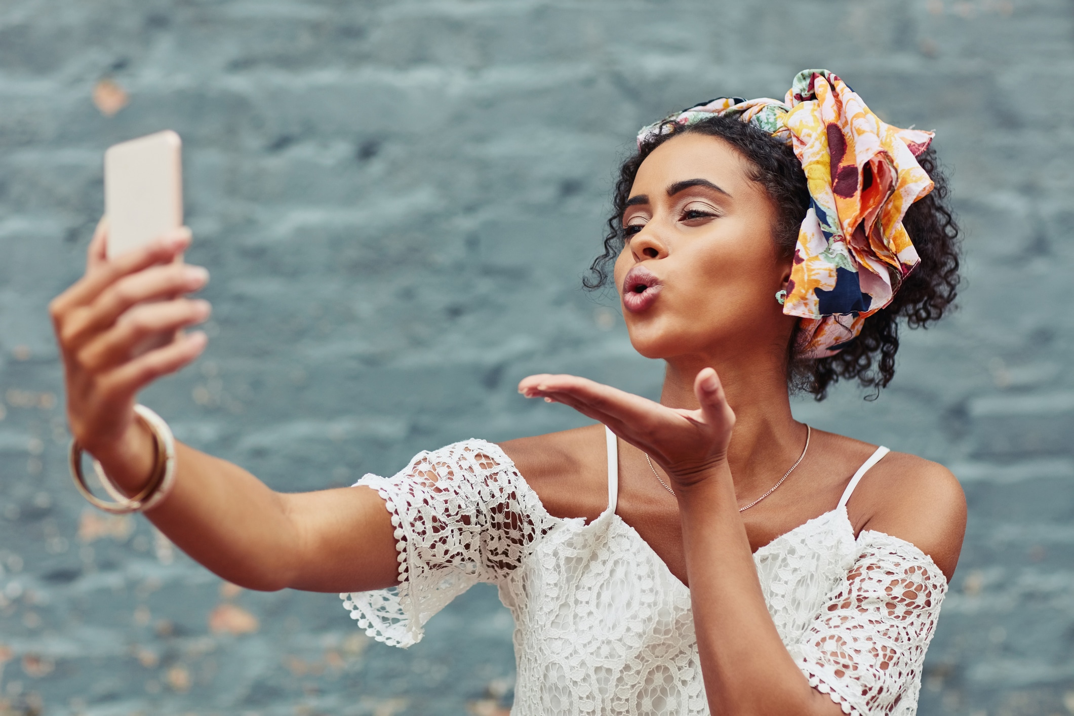  Mulher jovem com lenço na cabeça tirando selfie e enviando um beijo, vestindo blusa de renda branca