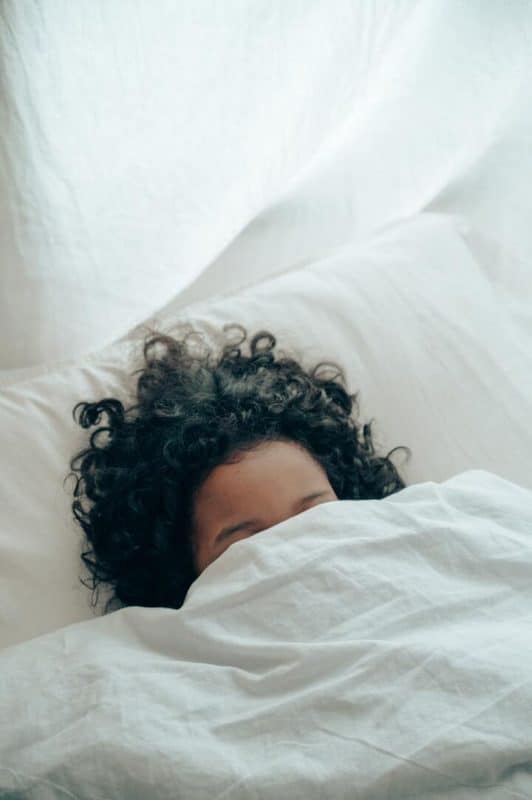 Mulher de cabelo cacheado com umectação noturna, deitada na cama, com um edredom branco cobrindo até os olhos