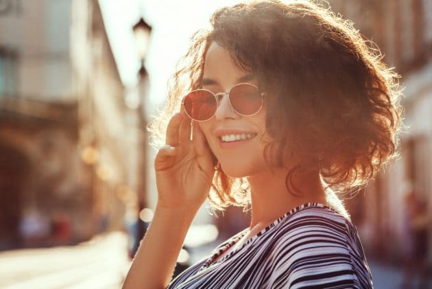 Mulher sorridente com óculos de sol redondos, estilo urbano ao pôr do sol