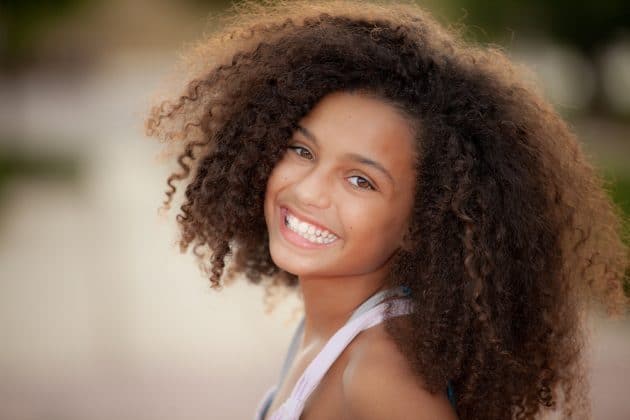 Menina negra sorridente, cabeça levantada, blusa branca e cabelos cacheados