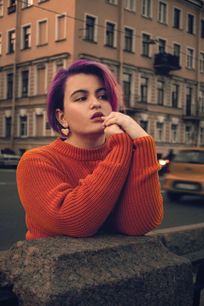 mulher branca com o cabelo curto pintado de roxo apoiada em um muro