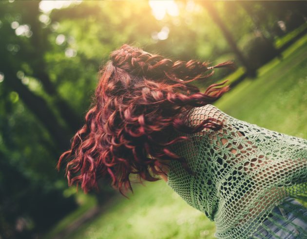 Mulher de costas, cabelo cacheado vermelho e blusa de renda, em uma floresta