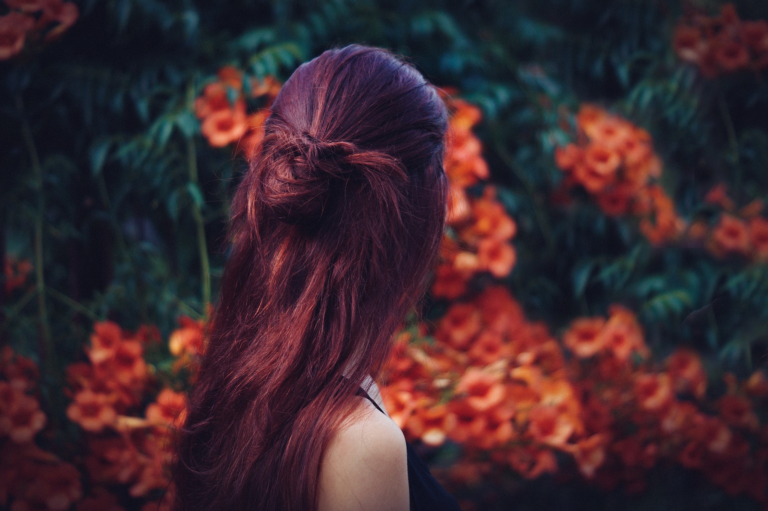 Mulher de costas, cabelo vermelho, com flores ao fundo na cor laranja, na floresta