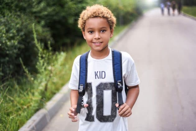 Menino negro sorridente, com blusa branca escrita "Kid 10", cabelo loiro e mochila azul