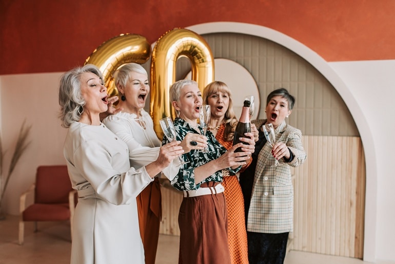  A imagem mostra com grupo de mulheres idosas (sendo 5, no total), com taças na mão, estourando um champanhe e brindando felizes