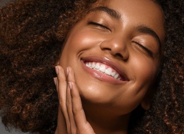 Mulher jovem, negra da pele mista e cabelo crespo, sorrindo com os olhos fechados, com a mão no rosto