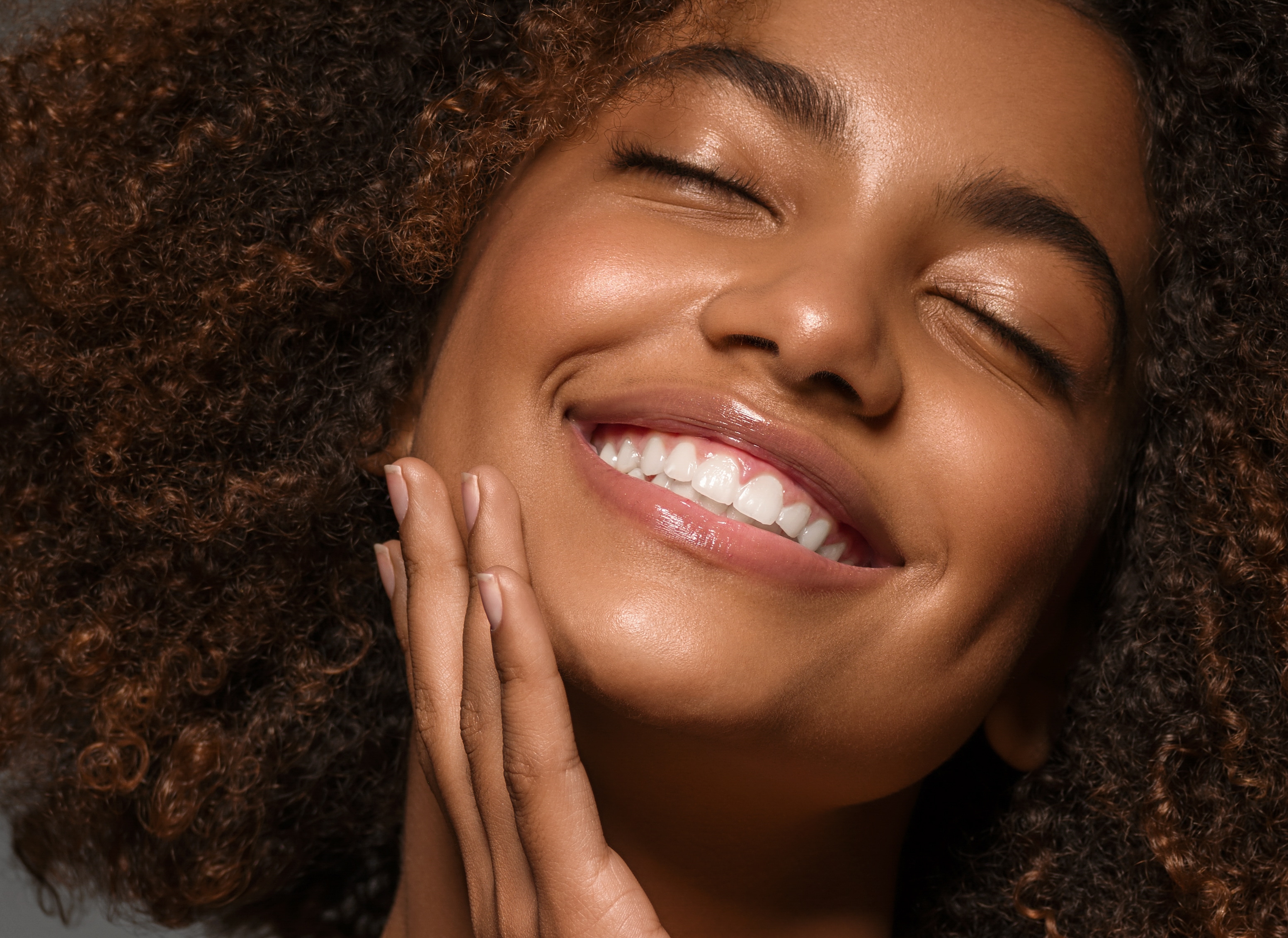  Mulher jovem, negra da pele mista e cabelo crespo, sorrindo com os olhos fechados, com a mão no rosto