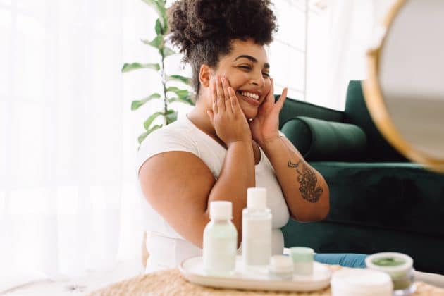 Mulher negra sorrindo com as mãos no rosto e produtos de skincare na mesa ao lado