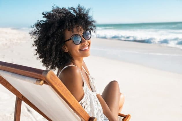 Mulher negra, de cabelo cacheado, na praia, usando óculos de sol