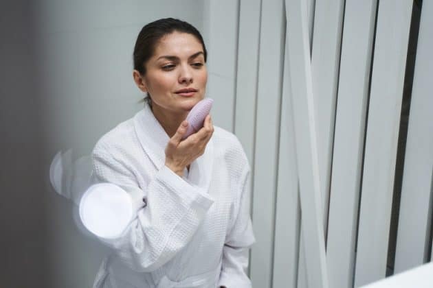 mulher branca, com roupão branco, fazendo skincare com esponja facial elétrica