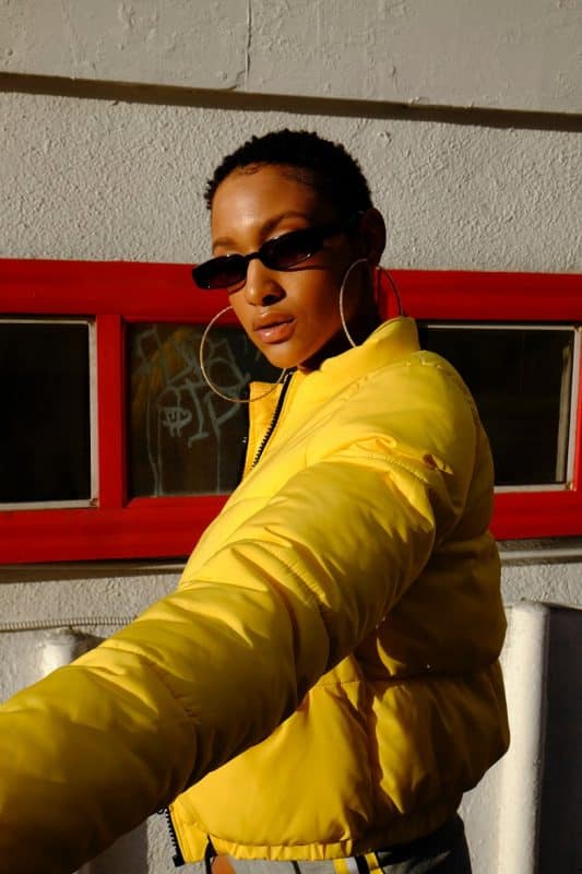 Mulher negra de cabelo crespo bem curtinho, blusa amarela, óculos de sol e maquiagem leve com gloss labial