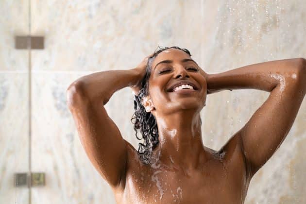 Mulher negra sorrindo enquanto hidrata o couro cabeludo ressecado no chuveiro
