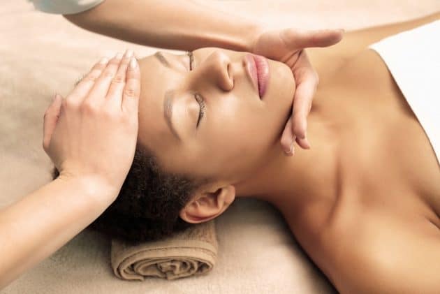 A imagem mostra uma mulher negra, deitada em uma maca de estética, com olhos fechados, enquanto um par de mãos brancas e femininas massageiam o seu rosto, para tratar a pele