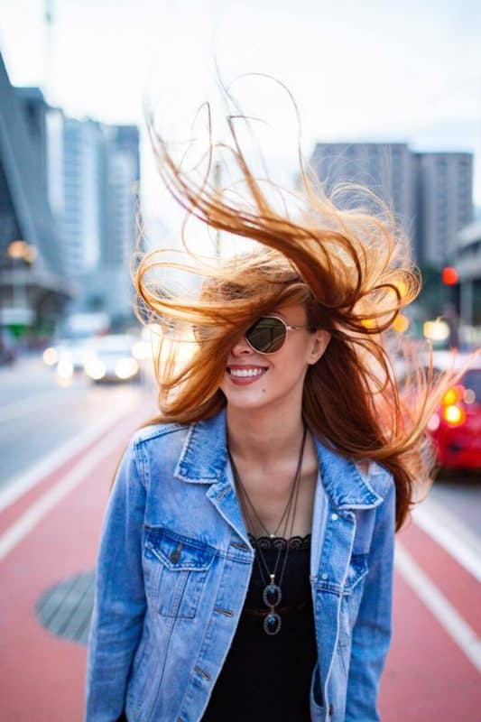 A imagem mostra uma mulher jovem, de cabelos longos, lisos e ruivos, esvoaçantes. Ela usa óculos de sol, jaqueta jeans e uma blusa preta por baixo, enquanto caminha sorridente pela rua