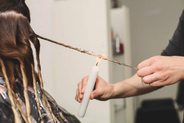 A imagem mostra as mãos de uma cabeleireira fazendo o processo de velaterapia capilar. O cabelo da cliente está dividido em mechas torcidinhas e a profissional está passando a chama da vela próximo aos fios de uma das mechas