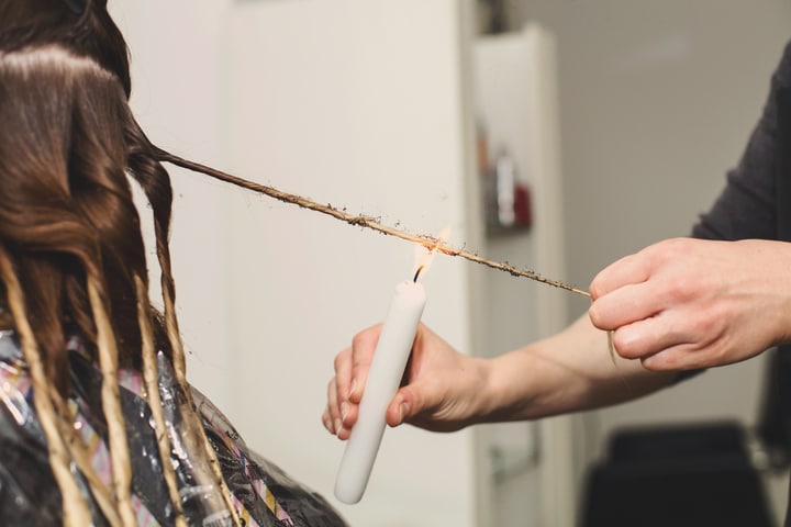  A imagem mostra as mãos de uma cabeleireira fazendo o processo de velaterapia capilar. O cabelo da cliente está dividido em mechas torcidinhas e a profissional está passando a chama da vela próximo aos fios de uma das mechas