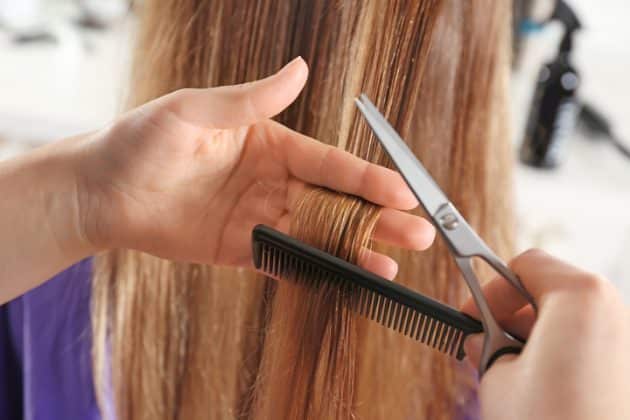 A imagem mostra uma cabeleireira fazendo um corte bordado em um cabelo loiro, liso e com pontas duplas