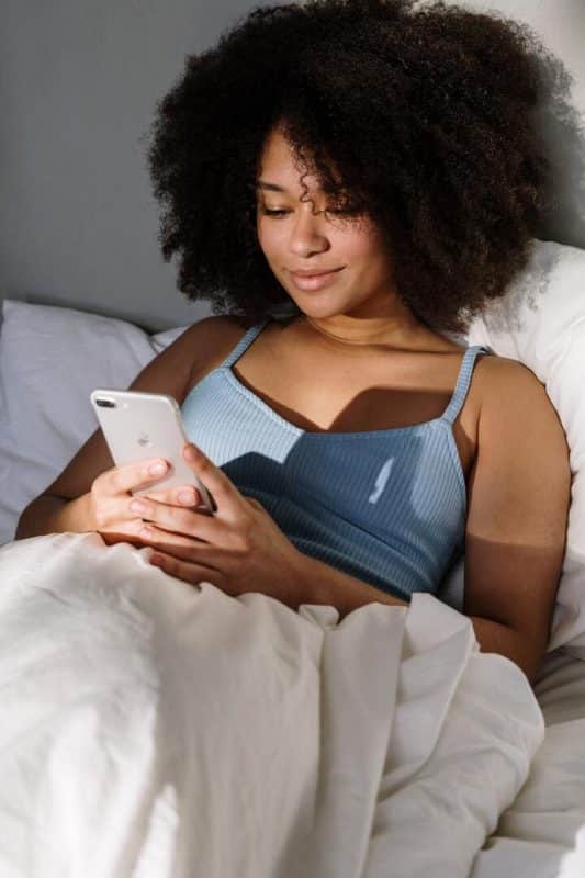 A imagem mostra uma mulher negra de cabelo crespo, preto, na altura dos ombros. Ela está na cama, encostada no travesseiro, enquanto digita no celular 