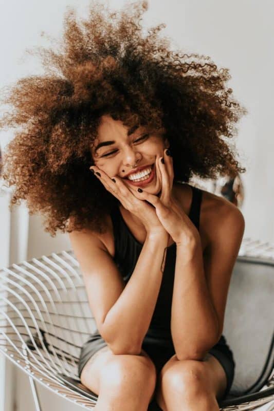 Mulher de cabelo crespo e longo, na cor castanho médio. Ela está sentada e sorrindo com as mãos no rosto