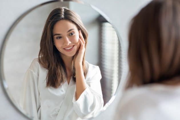 Mulher branca de cabelo liso castanho, se olhando no espelho e fazendo exercícios de yoga facial