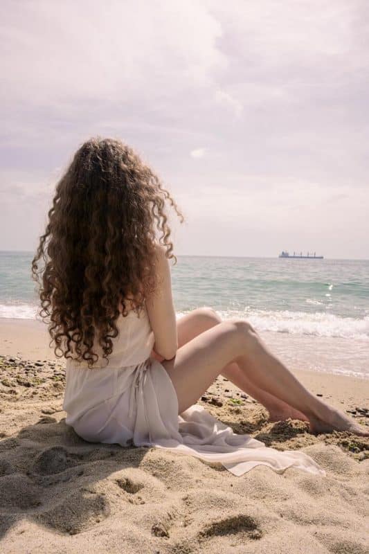 Mulher de cabelo cacheado castanho claro e longo. Ela está na praia, sentada na areia (de costas para a câmera), observando o mar