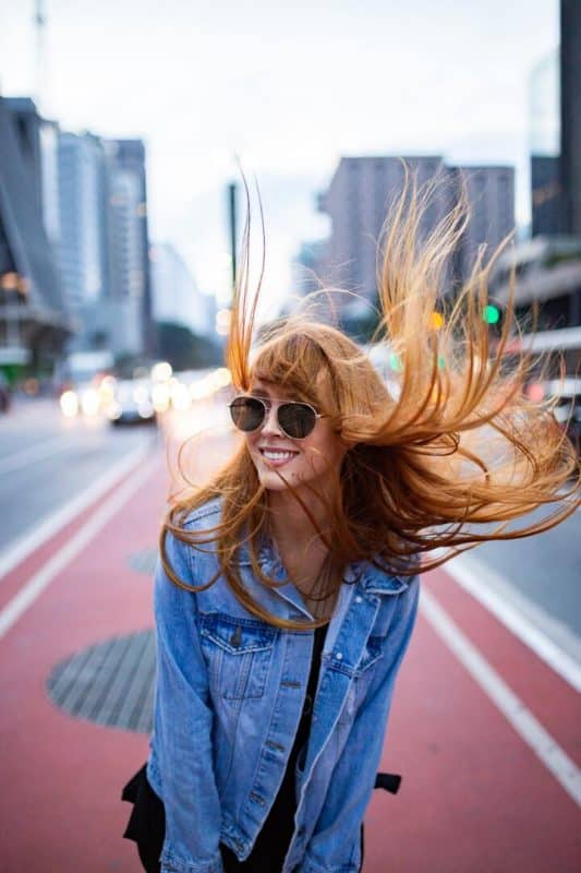 Mulher de cabelo ruivo liso e longo. Ela está de roupa preta, com uma jaqueta jeans azul por cima e óculos escuro.