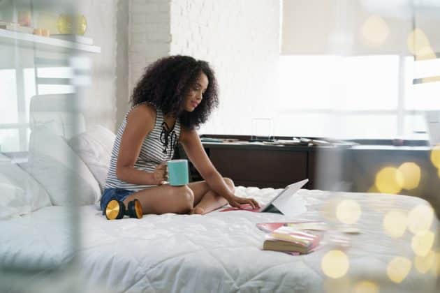 Mulher negra de cabelo cacheado, corte médio na altura dos ombros. Ela está sentada na cama, segurando uma xícara e mexendo no notebook