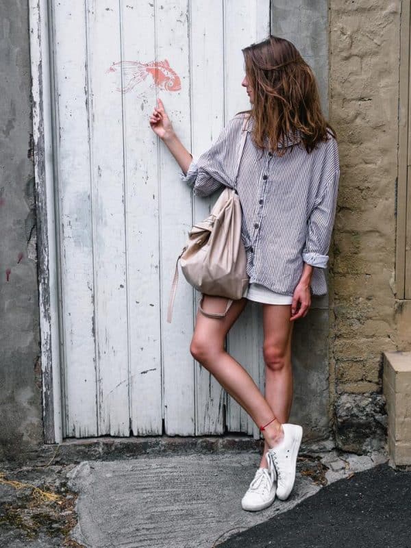 Mulher de cabelo liso, loiro, corte médio, usando uma camisa longa e quadriculada, uma mini saia branca, tênis branco e uma bolsa estilo bag, na cor bege