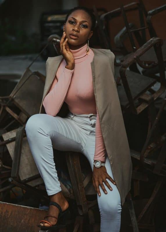 Mulher negra de cabelo preso, vestindo uma blusa rosê de manga longa e gola alta, calça jeans clara, um blazer bege, longo, jogado por cima e uma sandália preta, com uma tira laranja