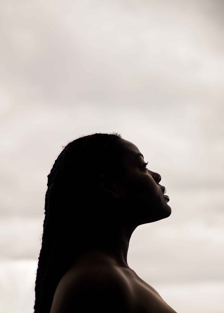  Mulher negra, de perfil, com cabelo trançado, olhando em direção ao horizonte, em forma de representação da solidão dessa mulher