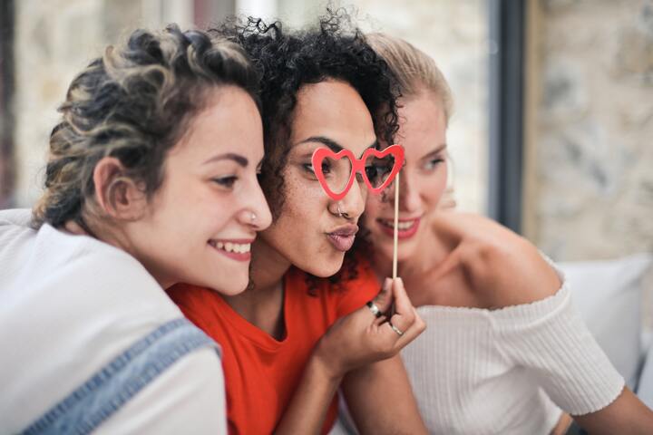  três mulheres posando para a foto, uma delas está com um óculos de coração vermelho.