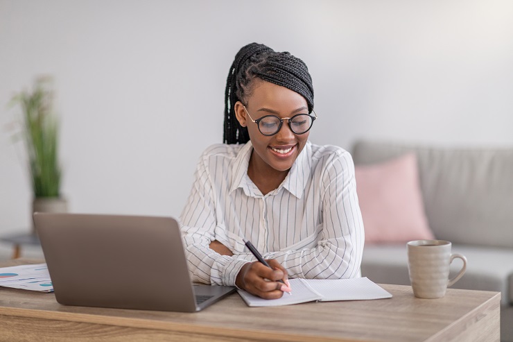 Mulher negra, com tranças box braids, sentada em uma mesa, com um notebook e uma folha de anotações, fazendo um curso de curta duração à distância